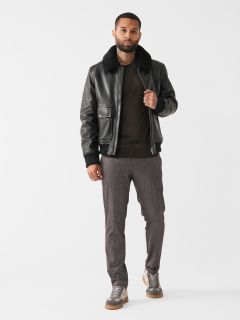 Ein Mann steht selbstbewusst vor einem weißen Hintergrund. Er trägt eine schwarze Lederjacke mit Pelzkragen, einen dunklen Pullover, eine graue Hose und graue Turnschuhe. Eine Hand steckt in der Jackentasche und er blickt direkt in die Kamera.