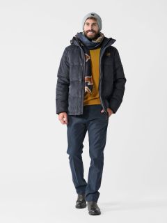 Ein Mann lächelt beim Gehen. Er trägt eine blaue Winterjacke, einen senffarbenen Pullover, einen Schal, blaue Hosen, schwarze Schuhe und eine hellgraue Mütze. Er hat einen Bart und posiert vor einem weißen Hintergrund.