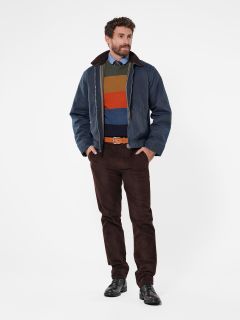 Ein Mann mit lockigem Haar, der eine blaue Jacke, einen bunt gestreiften Pullover, braune Hosen und schwarze Schuhe trägt, steht vor einem schlichten weißen Hintergrund. Er hat eine entspannte Haltung und die Hände in den Taschen.