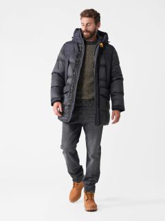 Ein bärtiger Mann in dunkler Pufferjacke, gemustertem Pullover und grauen Jeans geht selbstbewusst. Er trägt hellbraune Stiefel und an seiner Jacke hängt ein kleines orangefarbenes Accessoire. Der Hintergrund ist schlicht weiß.