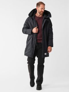 Ein Mann trägt einen langen schwarzen Mantel über einem kastanienbraunen Pullover und schwarzen Hosen sowie dunkle Schuhe. Er steht vor einem schlichten weißen Hintergrund, lächelt und blickt leicht nach unten.