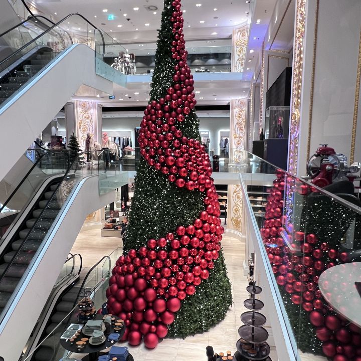 Ein großer Weihnachtsbaum in einem mehrstöckigen Einkaufszentrum ist mit roten Kugeln und einer Spirale aus roten Blumendekorationen geschmückt. Um den Baum herum sieht man Käufer und Rolltreppen, was zur festlichen Atmosphäre beiträgt.