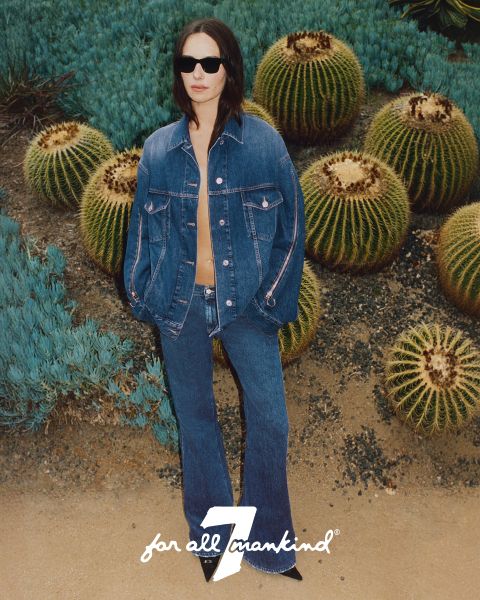 Eine Person in Jeansjacke und -hose steht zwischen Kakteen. Sie trägt eine Sonnenbrille und die Jacke ist leicht geöffnet. Der Boden ist sandig und im Hintergrund sind grüne Sukkulenten zu sehen. Unten ist das Logo „7 for all mankind“ zu sehen.