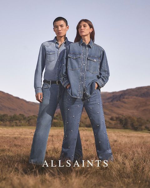 Zwei Personen stehen auf einem freien Feld und tragen Jeans-Outfits, die mit Nieten verziert sind. Die Person links trägt ein helles Jeanshemd und Jeans, während die Person rechts eine dunkle Jeansjacke und Jeans trägt. Unten steht „ALLSAINTS“.
