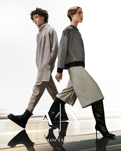 Zwei Models laufen nebeneinander, beide tragen übergroße Pullover und weite Hosen. Das Model links hat lockiges Haar und trägt Stiefel, während das Model rechts kurze Haare und glänzende Stiefel hat. Auf dem Bild ist das Logo von Armani Exchange zu sehen.