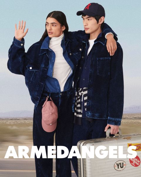 Zwei junge Erwachsene in Jeanskleidung stehen zusammen im Freien, einer mit erhobenem Arm, der andere mit einem Koffer, der mit Aufklebern bedeckt ist. Am unteren Rand steht in fetter weißer Schrift "ARMEDANGELS".