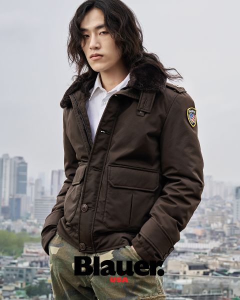 Eine Person mit langen Haaren, die eine braune Jacke und Camouflage-Hosen trägt, steht vor einer Stadtkulisse. Die Jacke hat einen Aufnäher auf der Schulter und unten ist das Wort „Blauer“ zu sehen. Der Himmel ist bewölkt.