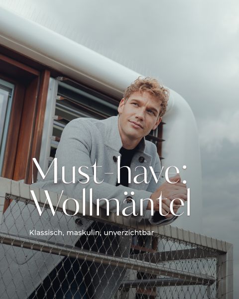 Ein Mann mit lockigem blondem Haar und einem hellgrauen Wollmantel lehnt sich an ein Metallgeländer im Freien, mit einem modernen Gebäude und einem bewölkten Himmel im Hintergrund. Der überlagerte Text lautet: "Must-have: Wollmäntel. Klassisch, maskulin, unverzichtbar.".