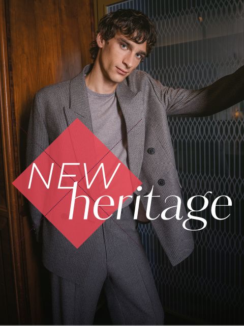 Ein Mann in einem grauen Anzug steht vor einer Holzwand, auf der der Schriftzug „NEW heritage“ in fetter Schrift zu sehen ist.