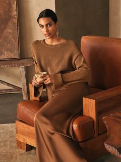 Eine Frau in einem braunen Pullover und einem langen braunen Rock sitzt auf einem Ledersessel und hält eine Tasse in der Hand; im Hintergrund ist eine Einrichtung in neutralen Farbtönen zu sehen.