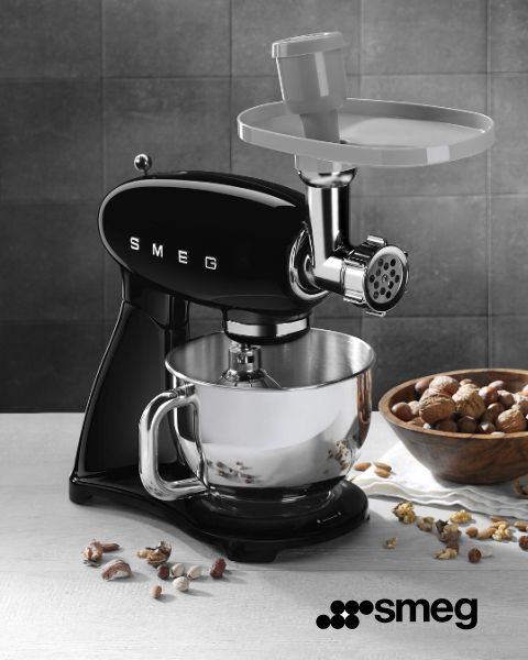 Eine schwarze Smeg-Küchenmaschine mit Fleischwolfaufsatz steht auf einer Küchentheke. Darunter befindet sich eine silberne Rührschüssel. Ganze und gehackte Nüsse liegen verstreut auf einer Holzfläche in der Nähe. In der unteren rechten Ecke ist das Smeg-Logo zu sehen.