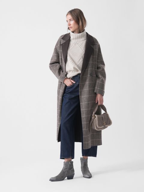 Eine Frau, die einen karierten Mantel, einen Pullover, eine dunkle Jeans und graue Stiefel trägt, hält eine kleine Handtasche in der Hand; sie steht mit dem Blick nach links vor einem schlichten weißen Hintergrund.