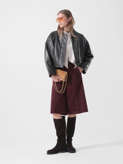 Eine Frau in einer schwarzen Lederjacke, einer burgunderroten Culotte, kniehohen Stiefeln und einer Sonnenbrille, die eine braune Handtasche in der Hand hält und vor einem schlichten weißen Hintergrund steht.