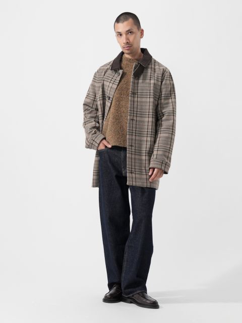 Ein Mann, der einen karierten Mantel, einen braunen Pullover, dunkle Jeans und schwarze Schuhe trägt, steht vor einem schlichten weißen Hintergrund.