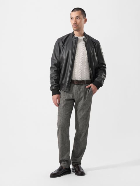 Ein Mann, der eine schwarze Lederjacke, einen weißen Pullover, eine graue Hose und dunkle Schuhe trägt, steht vor einem schlichten weißen Hintergrund und blickt leicht zur Seite.