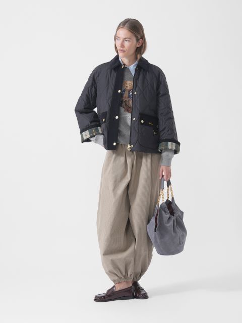 Eine Person in einer schwarzen Steppjacke, einem grauen Pullover, einer weiten beigen Hose und Slippern hält eine graue Tasche mit goldenen Kettengriffen in der Hand und steht vor einem weißen Hintergrund.