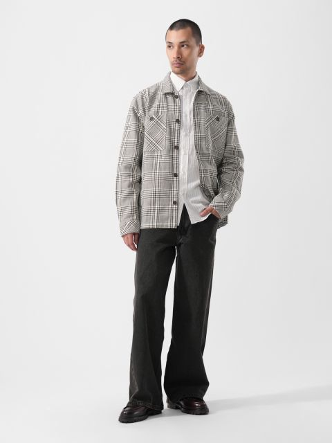 Vor einem schlichten weißen Hintergrund steht ein Mann, der eine karierte Jacke über einem weißen Hemd, eine weite schwarze Hose und schwarze Schuhe trägt.