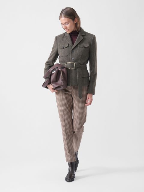 Eine Frau in einem Blazer mit Gürtel, einer karierten Hose und Stiefeln hält eine braune Tasche in der Hand, schaut nach unten und steht vor einem schlichten weißen Hintergrund.