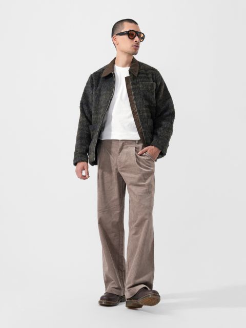 Full-body portrait of a man in a dark textured jacket, white tee, brown corduroy pants, and sunglasses against a plain light background.