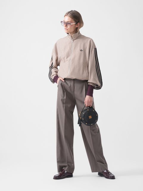 Eine Person mit Sonnenbrille trägt eine karierte Jacke, eine weite graue Hose und burgunderrote Schuhe und hält eine kleine runde schwarze Tasche vor einem schlichten weißen Hintergrund in der Hand.
