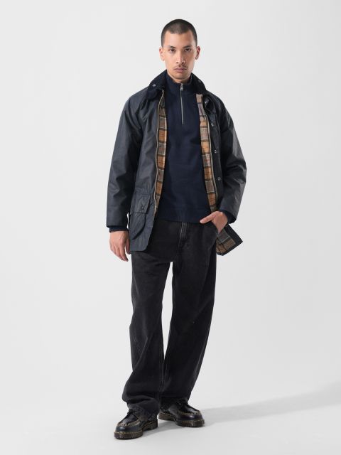Ein Mann steht in Freizeitkleidung vor einem schlichten weißen Hintergrund: dunkle Jacke mit kariertem Futter, marineblauer Pullover mit Reißverschluss, weite schwarze Jeans und schwarze Schuhe.