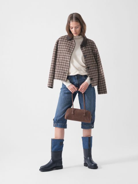Eine Frau trägt eine karierte Jacke über einem cremefarbenen Pullover, dazu hochgekrempelte Jeansshorts und marineblaue Stiefel; sie hält eine braune Handtasche in der Hand und steht vor einem schlichten Hintergrund.