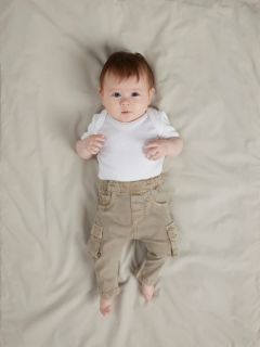 Ein lächelndes Baby liegt auf einer weißen Decke und trägt einen blauen Strickpullover, eine braune Cordhose mit Taschen und graue Socken. Das Baby sieht glücklich und entspannt aus und hat die Arme an den Ellbogen leicht angewinkelt.