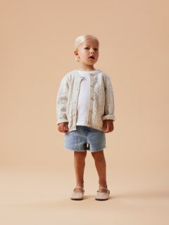 Ein lächelndes junges Mädchen in einem lavendelfarbenen Trainingsanzug steht vor einem grauen Hintergrund und trägt eine sehr große weiße Schleife auf dem Kopf.