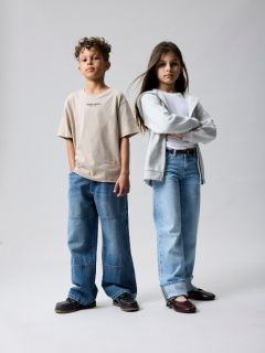 Zwei kleine Kinder stehen Händchen haltend vor einem einfarbig braunen Hintergrund. Ein Kind trägt eine rosafarbene Jacke und einen Rock, das andere eine blaue Jeans und ein blaues Hemd mit der Aufschrift "LIEBE IST KIND". Beide blicken neugierig nach oben.