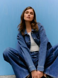 Eine Person mit langem, glattem blondem Haar und Pony trägt eine glänzende bordeauxfarbene Lederjacke und eine dazu passende Hose und lehnt an einer hellgrauen Steinmauer.