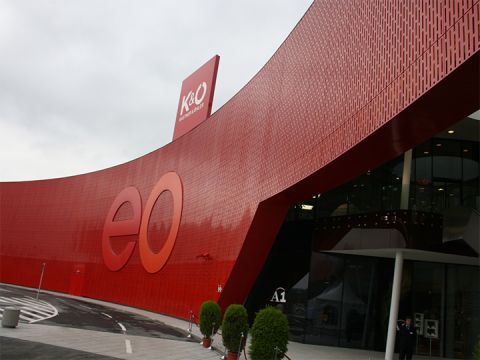 Ein modernes Gebäude mit einer großen, geschwungenen roten Fassade mit den Buchstaben „eo“. Auf dem Dach sind Schilder für K&O und Giga Sport zu sehen. Der Eingang ist aus Glas, daneben stehen Topfpflanzen, und im Vordergrund verläuft eine Kurve.