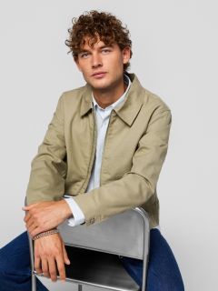 Ein junger Mann, der ein schwarzes T-Shirt mit der Aufschrift "Original Studio", eine silberne Halskette, ein silbernes Armband und eine creme-schwarz gestreifte Baseballkappe trägt, steht vor einem schlichten hellgrauen Hintergrund.