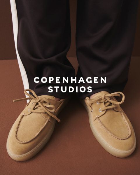 Eine Person in klobigen schwarzen Schnürstiefeln und dunklen Hosen steht auf einer weißen Fläche. Ein Teil des Bildes wird durch rosa und beigefarbenes, unscharfes Garn verdeckt. Der Text "Copenhagen Studios" erscheint über dem Foto.