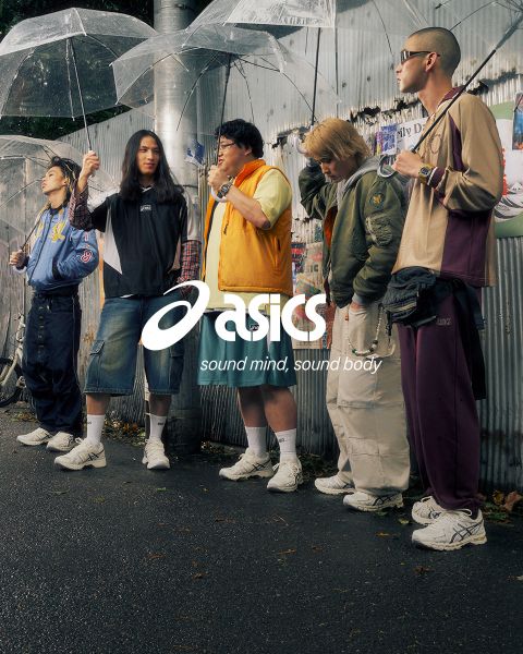 Zwei Personen in stylischen Outfits stehen mit Fahrrädern auf einer Kopfsteinpflasterstraße. Einer trägt ein blaues Hemd und weiße Hosen, der andere eine dunkle Jacke und Hosen. Die Szene ist sonnig und im Hintergrund sind alte Gebäude zu sehen. Das Asics-Logo ist sichtbar.