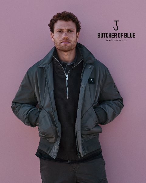 Ein Mann mit lockigem Haar trägt eine dunkelgrüne Jacke über einem Pullover mit Reißverschluss und steht vor einem rosa Hintergrund. Das Logo "Butcher of Blue Quality Clothing Co" ist in der oberen rechten Ecke zu sehen.