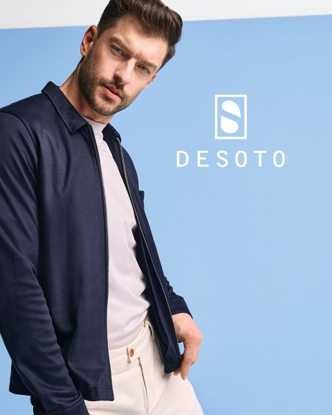 Ein Mann mit einem langärmeligen, gemusterten Hemd und einer dunklen Hose steht vor einem grauen Hintergrund. Er blickt mit den Händen in den Taschen zur Seite. Der Schriftzug "DESOTO" und das DESOTO-Logo sind am unteren Rand zu sehen.