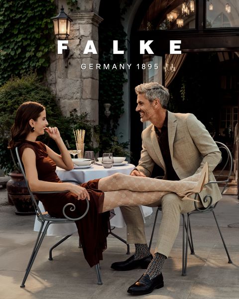 Ein Modell in einem kastanienbraunen Blazer, braunen Handschuhen, einer grünen Strumpfhose, einem grauen Rock mit hohen Schlitzen und braunen Ankle Boots posiert vor einem geometrischen Hintergrund. Der Text lautet "FALKE GERMANY 1895".