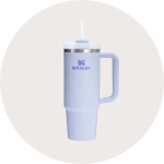 Ein cremefarbener Stanley-Becher mit Henkel, Edelstahlrand und einem Deckel mit Strohhalm, dargestellt auf einem hellgrauen, runden Hintergrund.