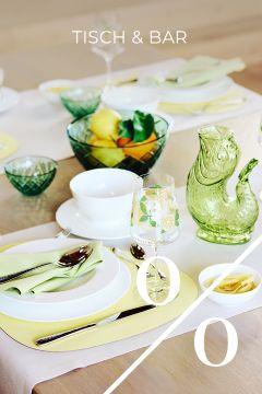 Ein für eine Mahlzeit gedeckter Tisch mit gelben Tischsets, weißen Tellern, grünen Servietten, Gläsern, einem grünen fischförmigen Krug, einer Schale mit Obst und dem Text "ESSEN & TRINKEN" am oberen Rand. Eine diagonale Linie mit "0%" überlagert das Bild.