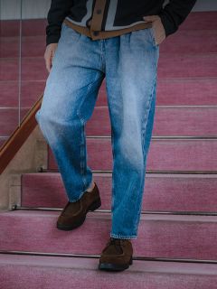 Ein Mann in weißem T-Shirt, dunkler Jeansjacke und Jeans steht an einem Geländer, das an eine Betonmauer und einen Zaun gelehnt ist, darüber eine gelbe Markise.