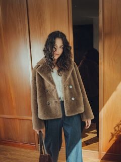 Eine Frau mit lockigem Haar, die einen braunen Kunstpelzmantel, eine weiße Bluse und Jeans mit weiten Beinen trägt, steht in einem Innenraum mit einer braunen Tasche in der Hand, mit Holzpaneelen und Sonnenlicht im Hintergrund.