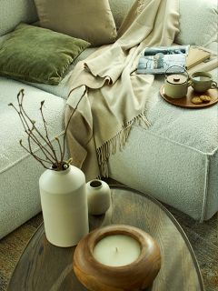 Gemütliche Wohnzimmerszene mit einem weichen, hellen Sofa, grünen und beigen Kissen, einer beigen Überwurfdecke, einem Tablett mit Tee und Keksen, einer Zeitschrift, Büchern und einem hölzernen Couchtisch mit einer Vase, einer Kerze und dekorativen Zweigen.