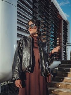 Eine Frau mit einer großen Sonnenbrille und einer dunklen Lederjacke lehnt an einer Wand, die Hände hinter dem Kopf verschränkt, und posiert selbstbewusst im Sonnenlicht.