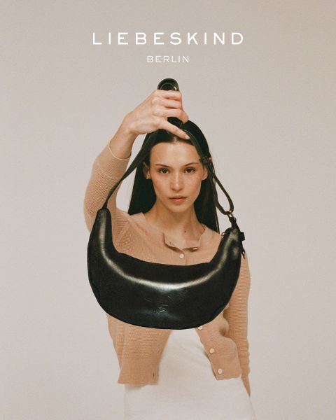 Eine lächelnde Frau in einem schwarzen langärmeligen Hemd und Jeans trägt eine hellbraune Tasche über der Schulter. Der Text "Liebeskind Berlin" erscheint am unteren Rand des Bildes.