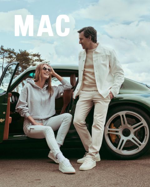 Eine Frau in einer Pelzjacke und weiten Hosen steht neben einem Mann in einer beigen Jacke und khakifarbenen Hosen. Beide posieren selbstbewusst im Freien vor einem steinernen Geländer unter blauem Himmel. Das Wort "MAC" erscheint am unteren Rand.