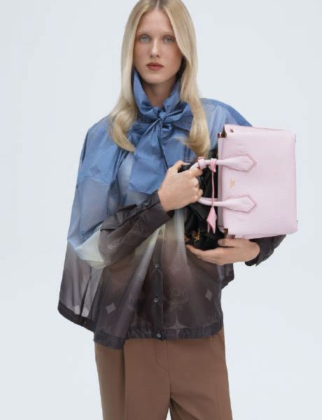 Eine grüne Designer-Handtasche mit dezentem Monogrammmuster ruht auf einer grauen Fläche, mit einem blauen geometrischen Objekt im unteren Vordergrund und einem Schattenwurf im Hintergrund.