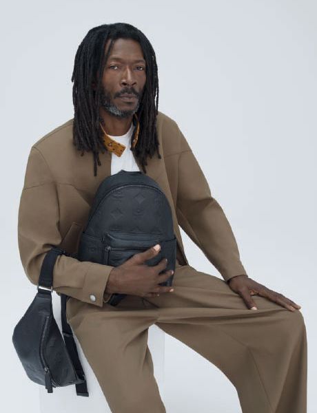 Ein grüner Designerrucksack und eine dazu passende Umhängetasche stehen auf einem Stapel orangefarbener und brauner Kisten vor einer beigefarbenen Strukturwand und einem gefliesten Boden.