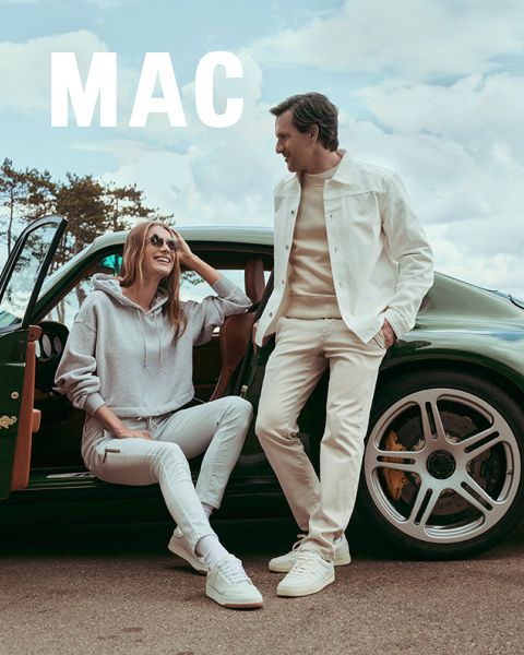 Eine Frau in einer Pelzjacke und weiten Hosen steht neben einem Mann in einer beigen Jacke und khakifarbenen Hosen. Beide posieren selbstbewusst im Freien vor einem steinernen Geländer unter blauem Himmel. Das Wort "MAC" erscheint am unteren Rand.