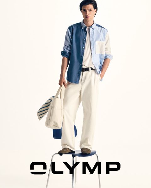 Ein Mann in Anzughose, kariertem Hemd und Krawatte mit einem über die Schultern drapierten Pullover steht vor einem dunkelblauen Hintergrund, neben dem das Wort "OLYMP" zu lesen ist.