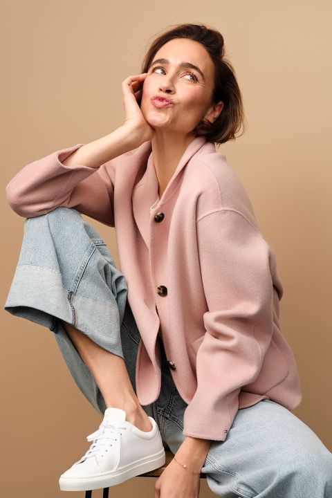 Eine Frau in rosa Strickjacke und Jeans sitzt verspielt auf einem Hocker und stützt ihr Kinn auf ihr Knie. Sie neigt den Kopf und zieht einen verspielten Schmollmund, die Hand ruht auf ihrer Wange. Sie trägt weiße Turnschuhe und posiert vor einem beigen Hintergrund.